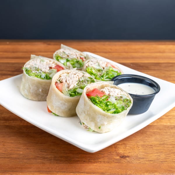 Several chicken salad wraps with lettuce and tomato on a white plate with ranch dressing.