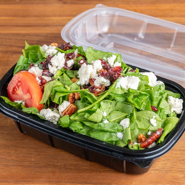 Spinach salad with feta, cranberries, pecans, and tomato in a black takeout container with an open lid.