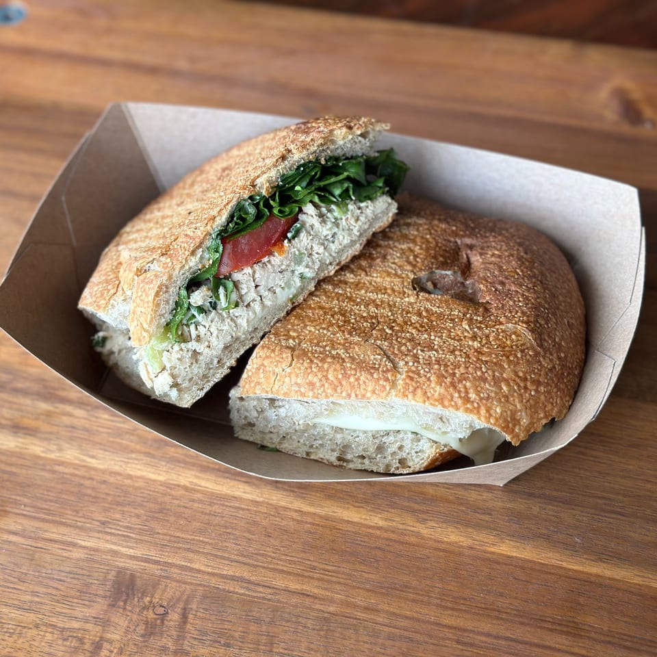 A panini sandwich with tuna salad, lettuce, tomato, and cheese in a paper tray on a wooden table.