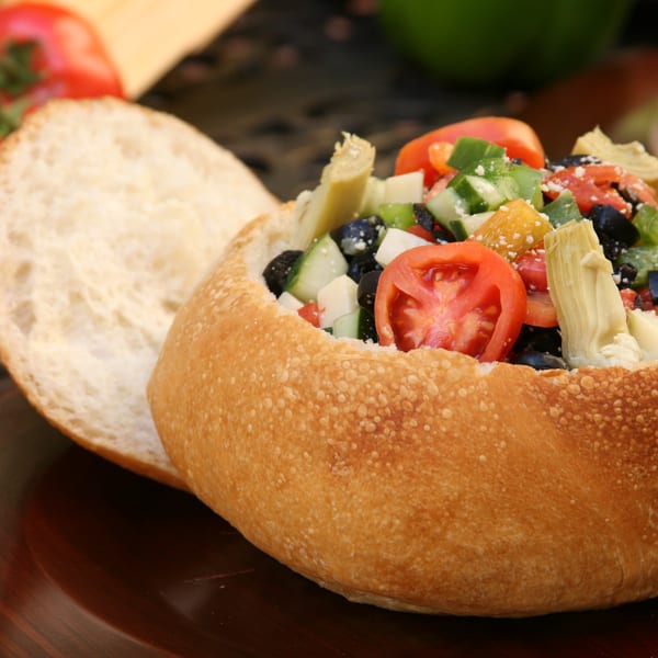 A bread bowl filled with a colorful salad of tomatoes, cucumbers, olives, and artichoke hearts.