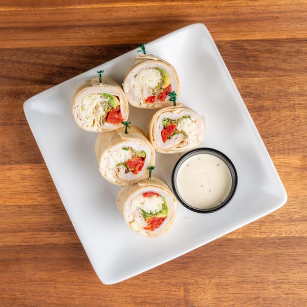 Five turkey and avocado pinwheel wraps with sun-dried tomatoes and dipping sauce on a white plate.