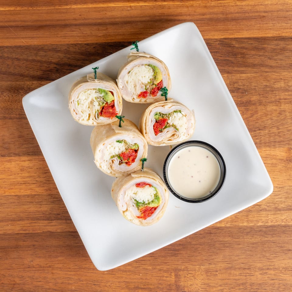 Five turkey and avocado pinwheel wraps with sun-dried tomatoes and dipping sauce on a white plate.